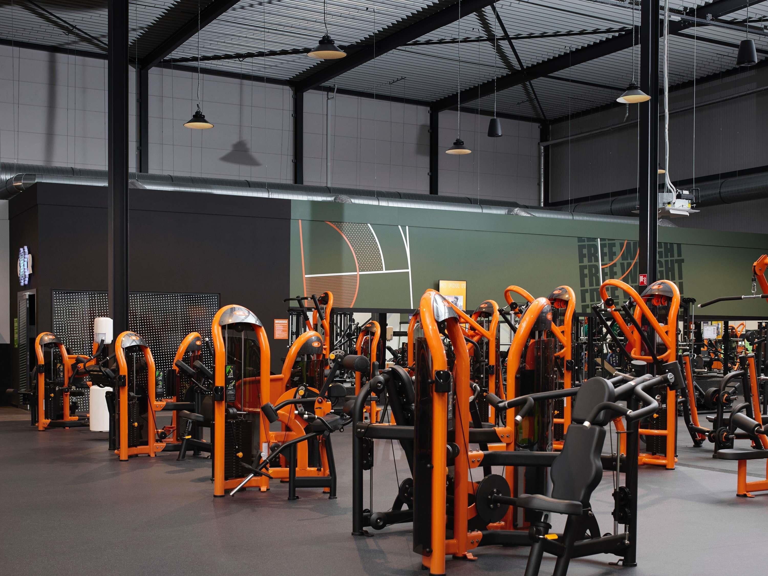 Un gimnasio con diversas máquinas de ejercicio, un espejo grande y un techo alto con vigas a la vista.