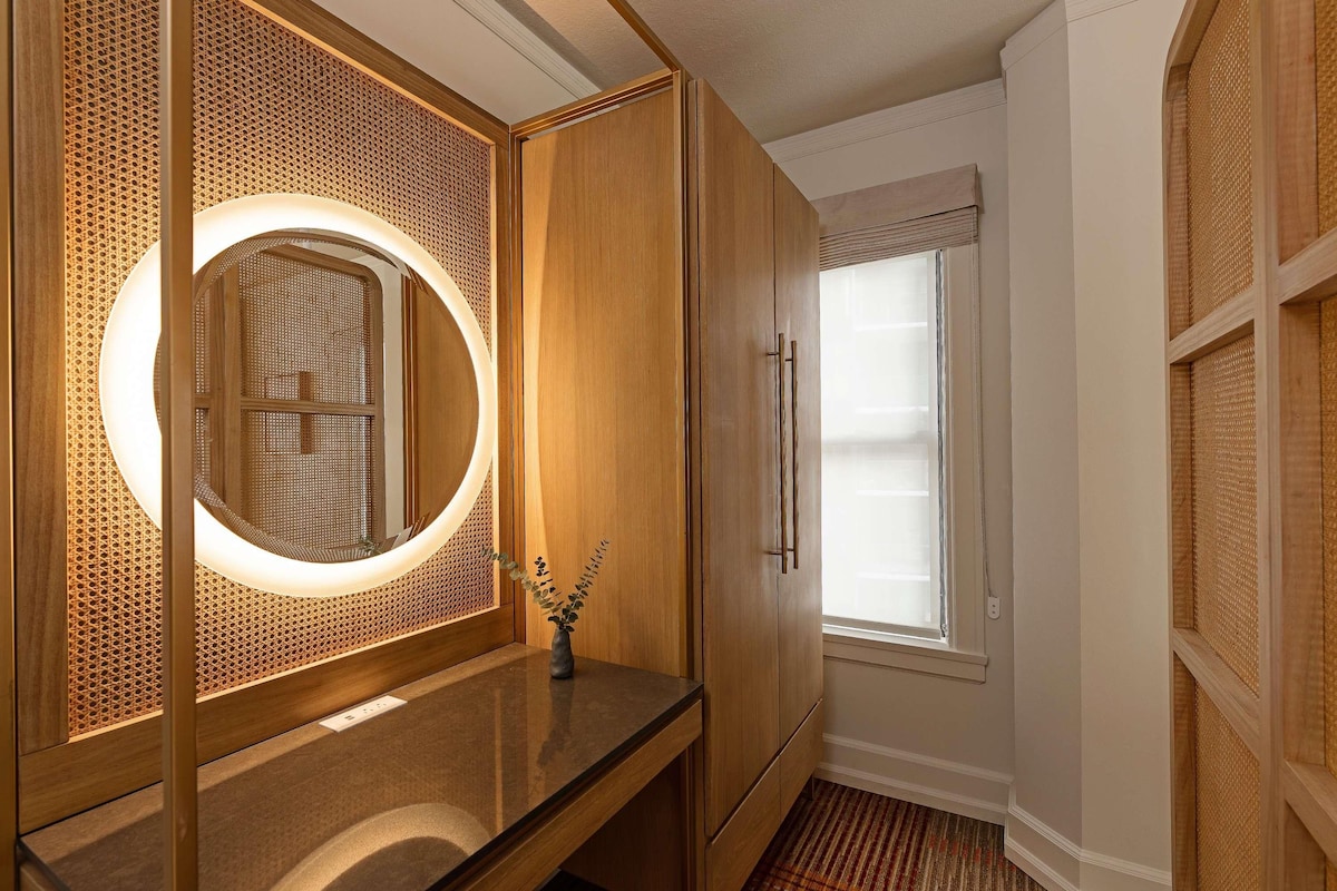 A bathroom with a wooden vanity, a round mirror with a light, and a window with blinds.