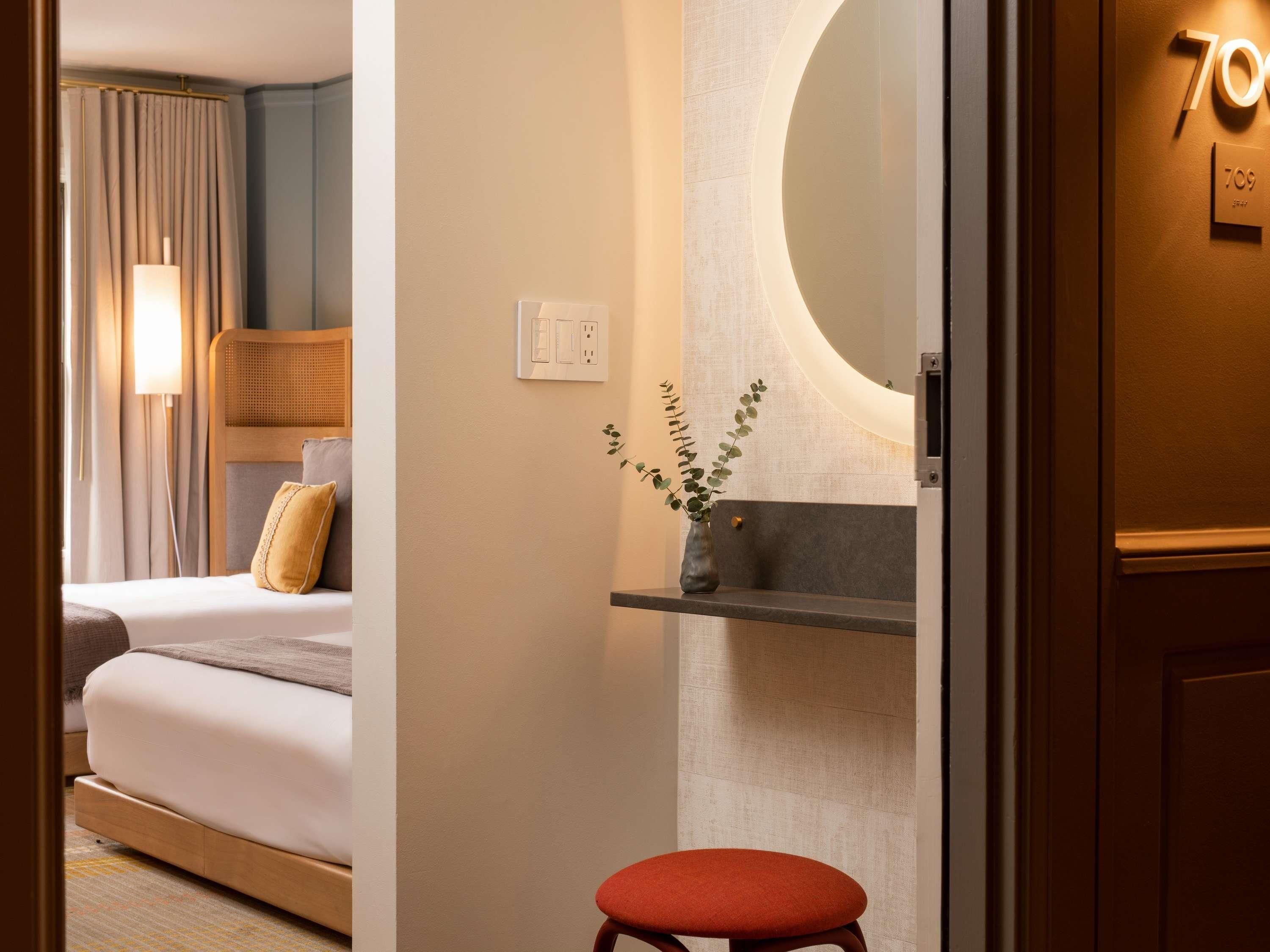 A modern hotel room with a bed, a bedside table, a mirror, and a small stool.