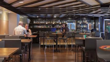 Zona de bar moderna con suelos de madera, taburetes de metal y una variedad de botellas de licor en estanterías.