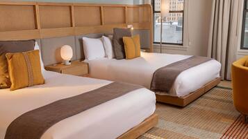 Ein Hotelzimmer mit zwei Betten, einem Nachttisch, einer Lampe und Blick auf die Stadt durch große Fenster.