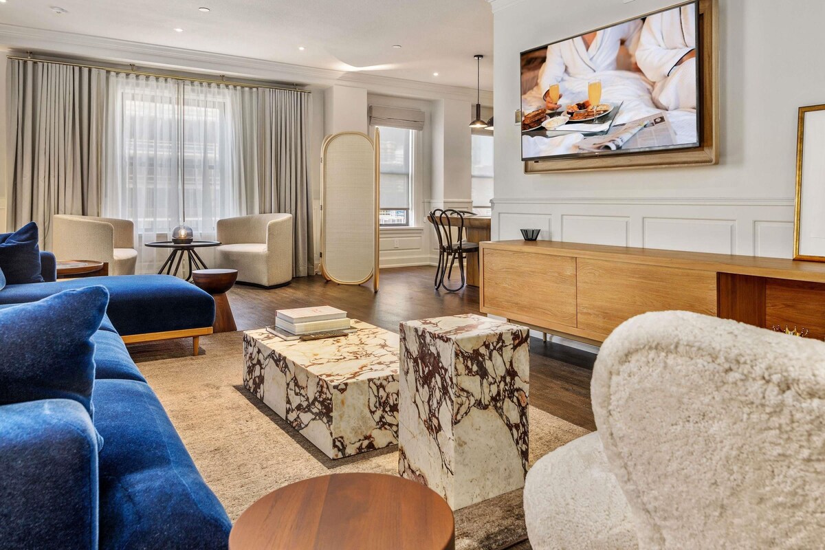 A modern living room with a blue sofa, a white armchair, marble coffee tables, and a flat-screen TV mounted on the wall.