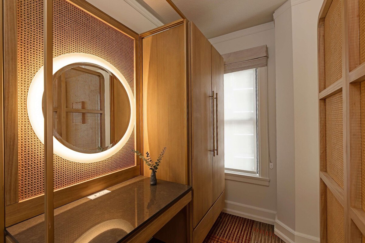 A bathroom with a wooden vanity, a round mirror with a light, and a window with blinds.