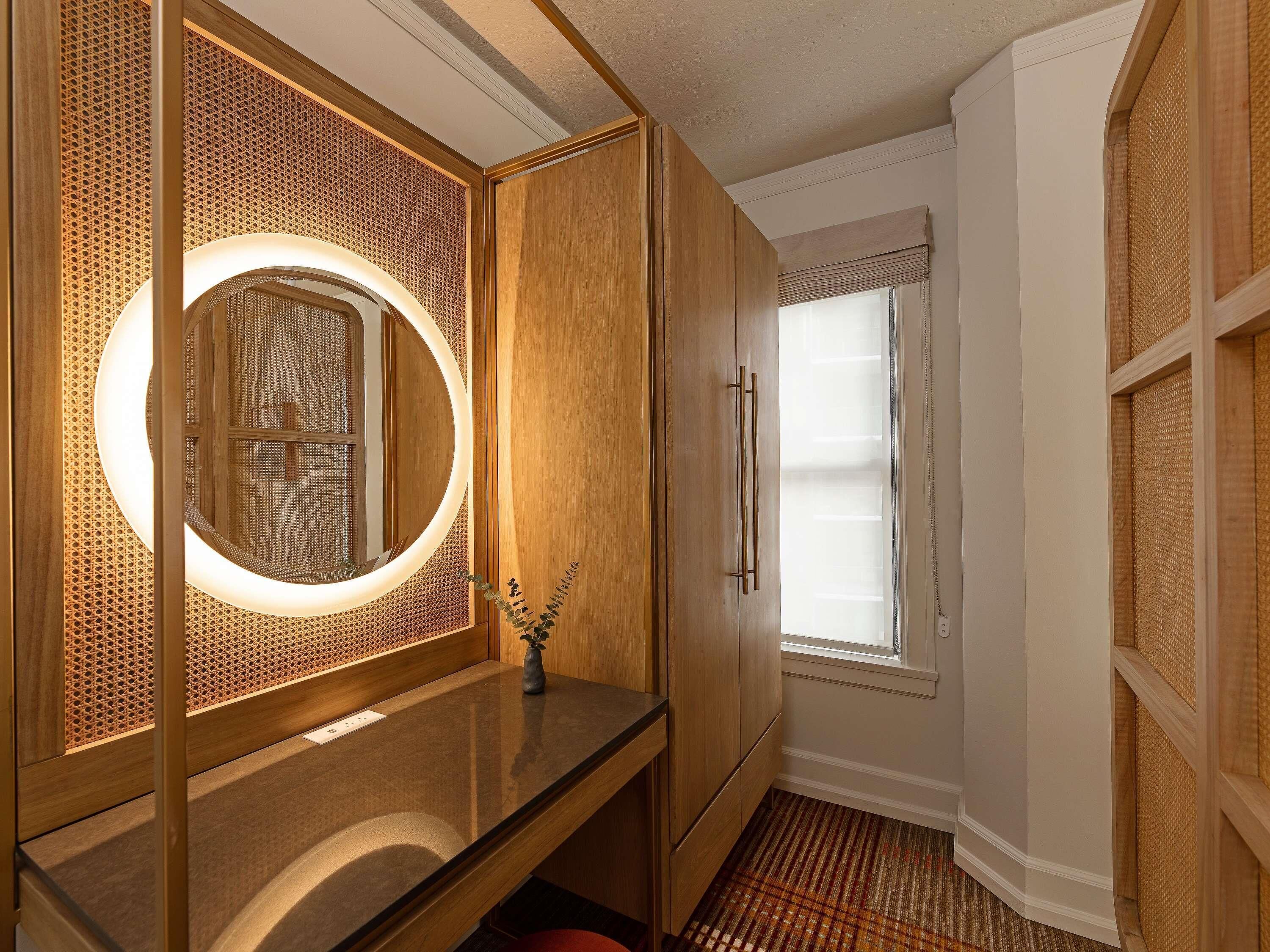Un baño con tocador de madera, un espejo redondo con luz y una ventana con persianas.