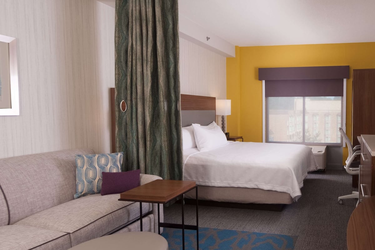 A hotel room with a bed, a sofa, a small table, a desk with a computer, and a window with a view of buildings.