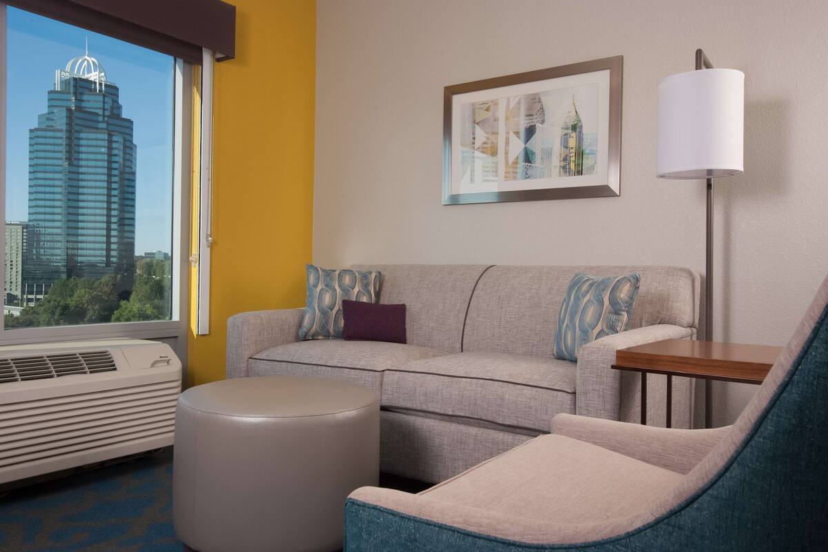 A hotel room with a grey sofa, a blue armchair, a small wooden table, a standing lamp, and a framed cityscape picture on the wall.