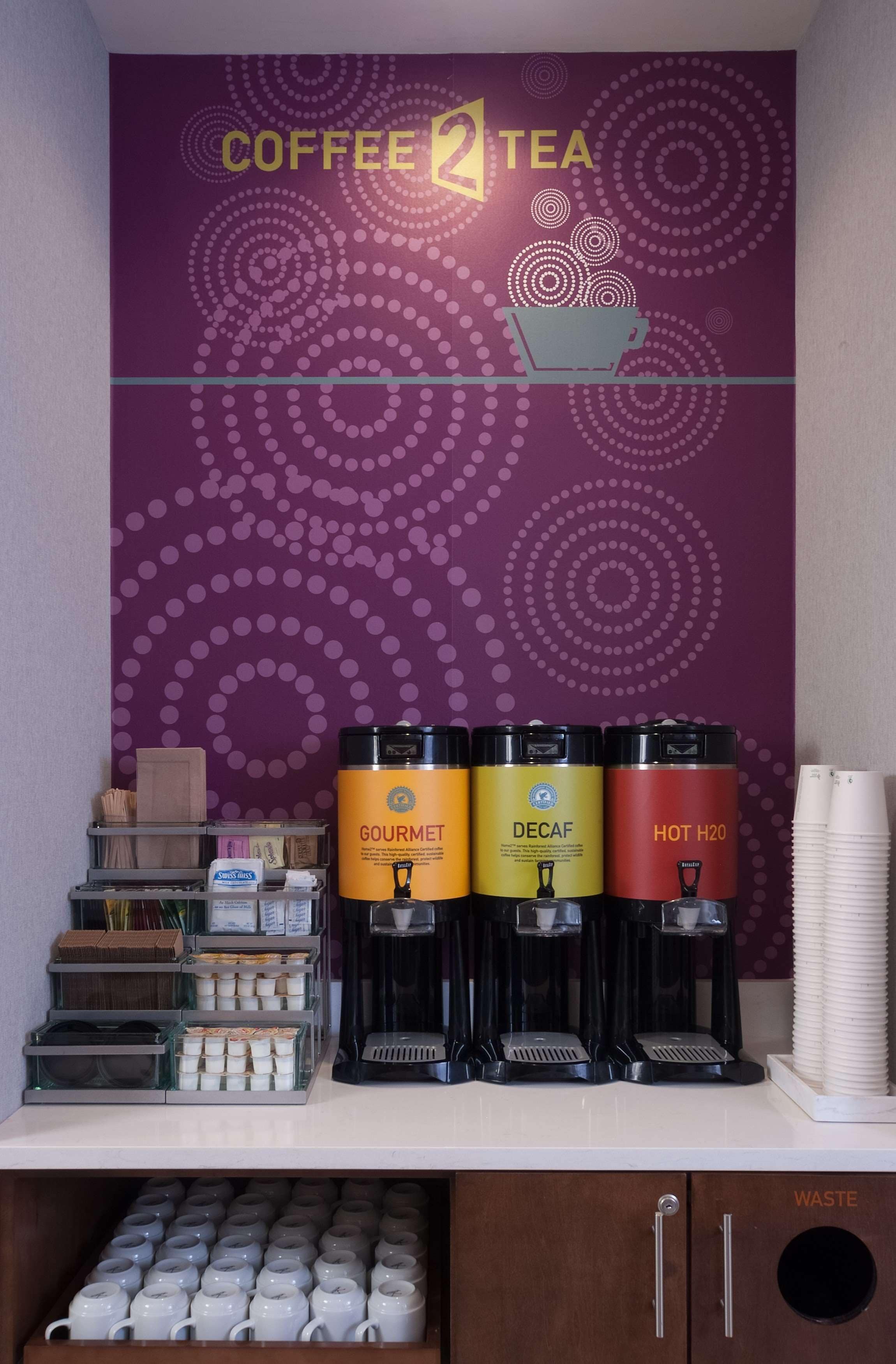 A coffee and tea station with three dispensers labeled 'Gourmet,' 'Decaf,' and 'Hot H2O.' There are stacks of disposable cups and a shelf with various items.
