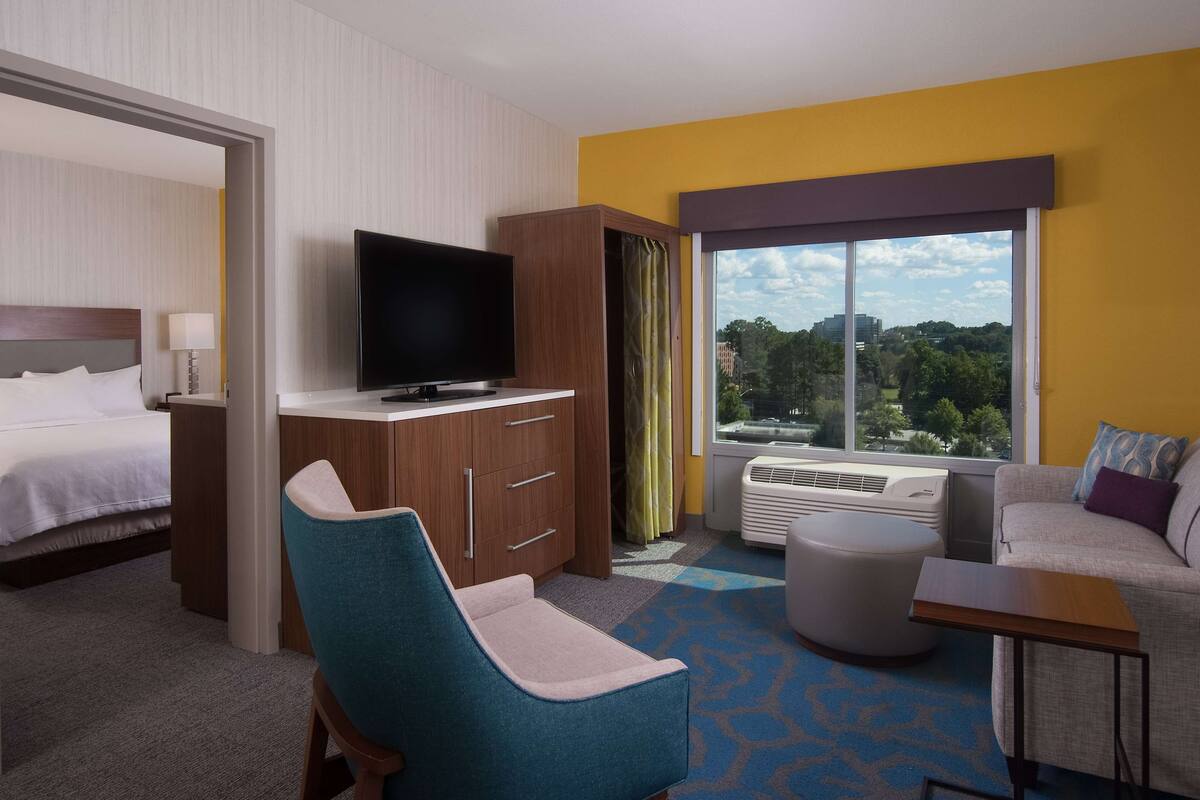A hotel room with a bed, a TV, a desk, a chair, a sofa, and a large window with a city view.
