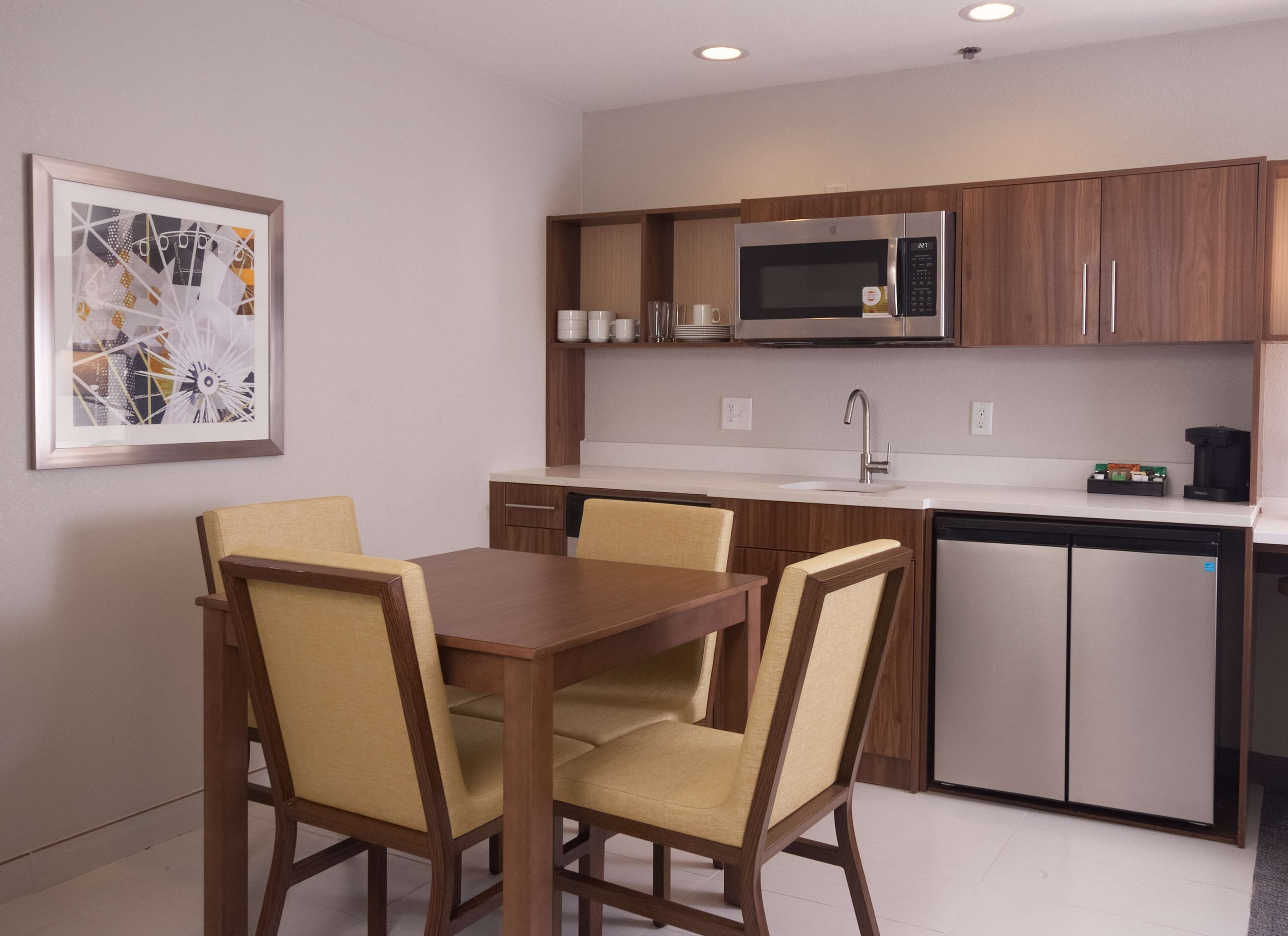 A compact kitchen with a microwave, sink, and dishwasher. There is a dining table with chairs and a framed picture on the wall.