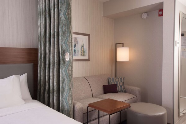 A hotel room with a bed, a sofa, a small table, a lamp, and a framed picture on the wall. - Home2 Suites by Hilton Atlanta Perimeter Center (Atlanta)
