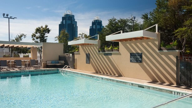 A swimming pool with a view of tall buildings in the background, a covered seating area, and a sign indicating a maximum bath load of 26 people. - Home2 Suites by Hilton Atlanta Perimeter Center (Atlanta)