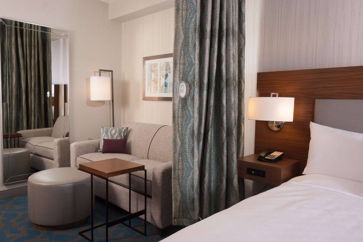 A hotel room with a bed, a sofa, a small table, a nightstand with a phone, and a mirror.