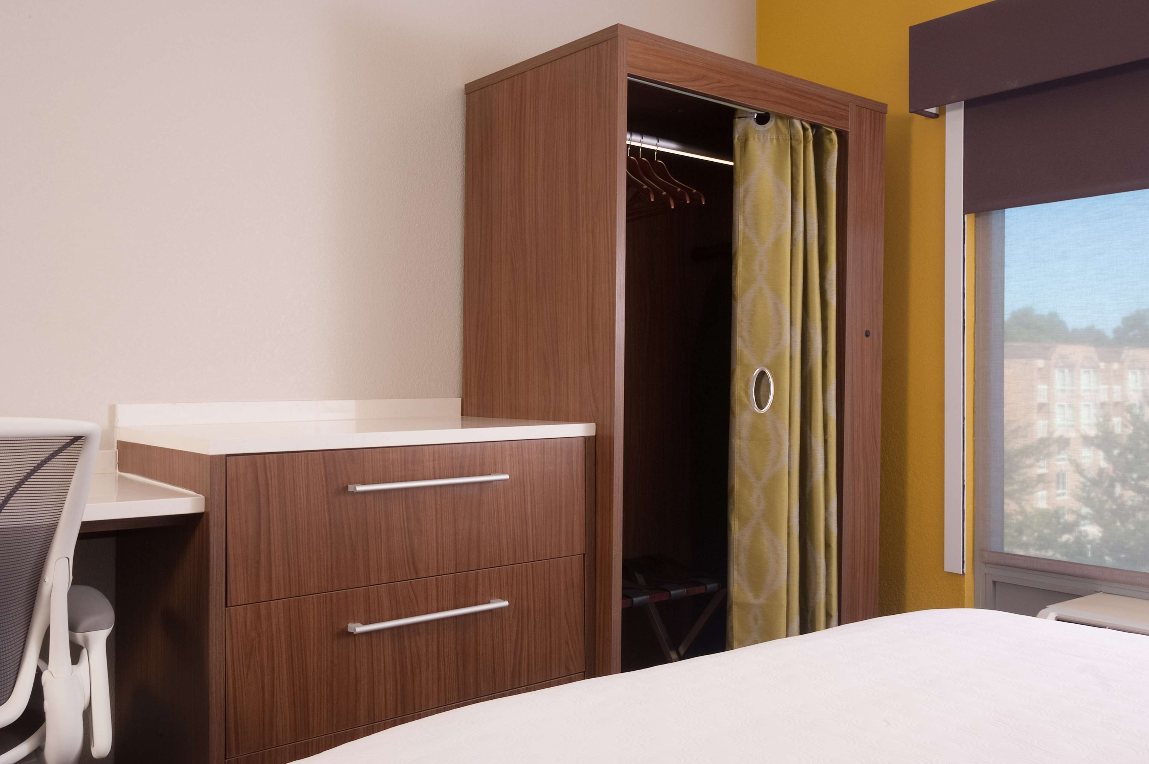 Une chambre d’hôtel avec une armoire en bois, un lit, un bureau avec une chaise et une fenêtre donnant sur des immeubles.