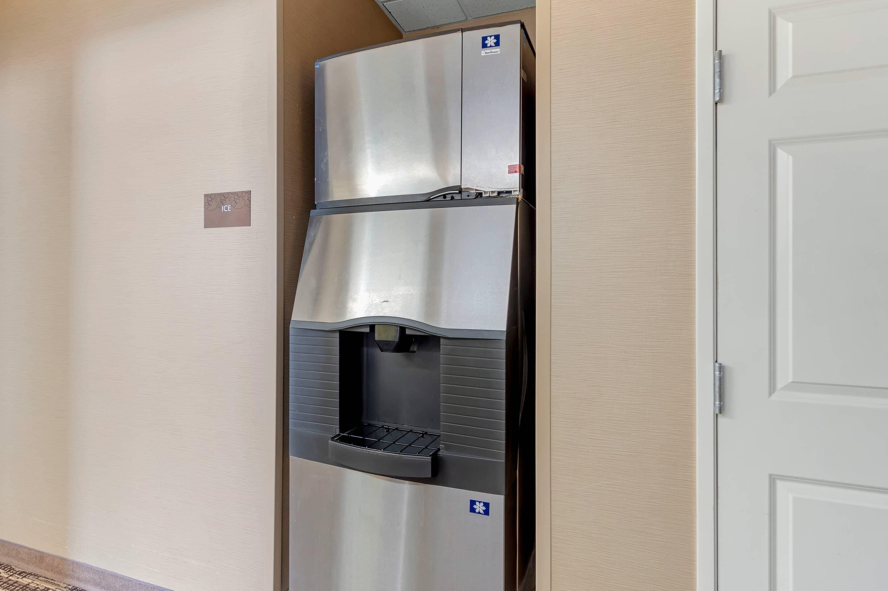 Um bebedouro de aço inoxidável com área para servir bebidas, localizado em um corredor com paredes bege e uma porta branca.