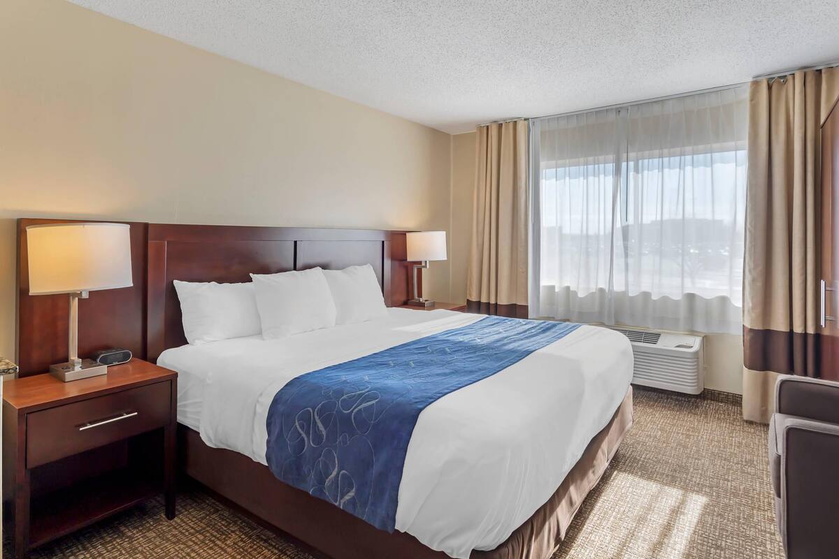 A hotel room with a large bed, two bedside tables with lamps, a desk with a chair, and a large window with curtains.
