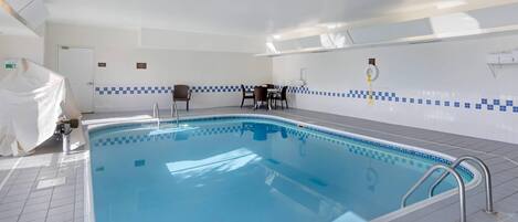 An indoor swimming pool with a tiled floor, a poolside table, and chairs.