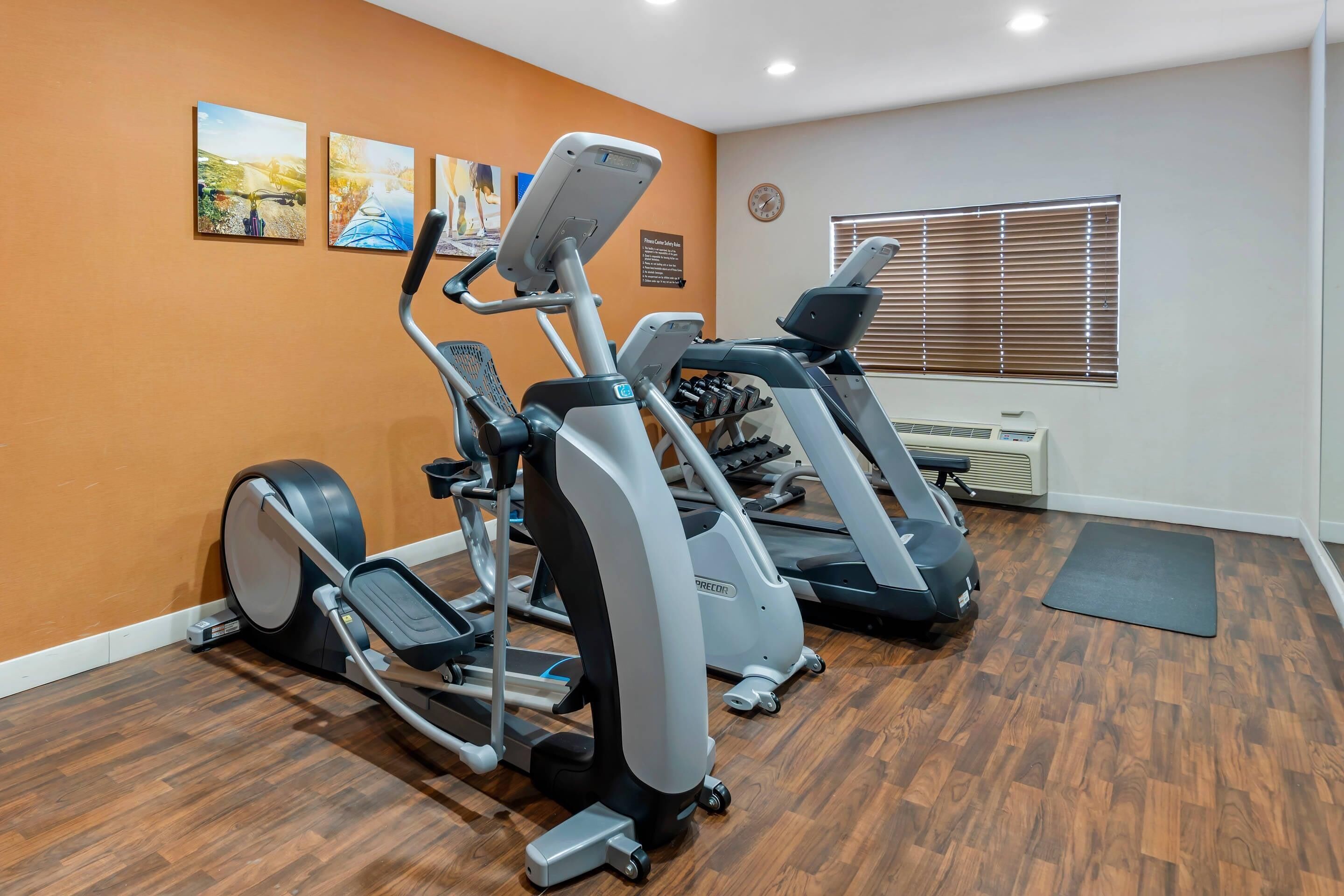 A gym with exercise equipment, wooden flooring, and a window with blinds.