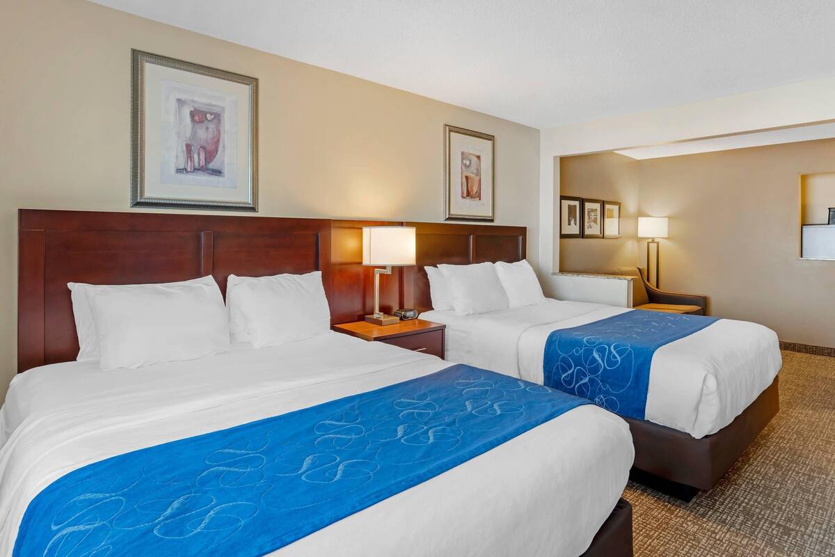 A hotel room with two beds, a wooden headboard, a nightstand with a lamp, and framed artwork on the walls.