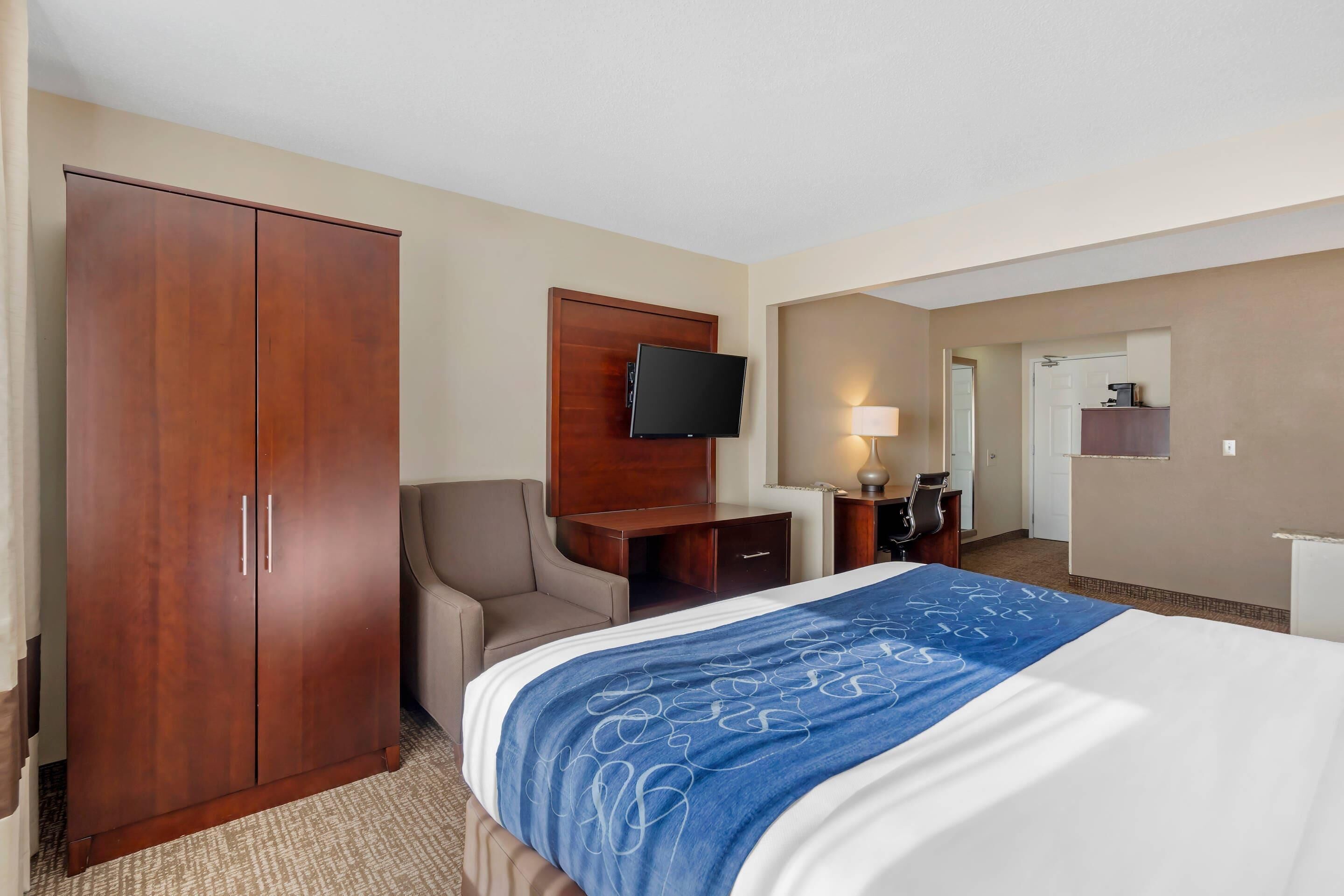 A hotel room with a large bed, a wardrobe, a desk with a flat-screen TV, a chair, a desk lamp, and a small kitchenette area.