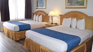 Habitación de hotel con dos camas, cada una con cabecero de madera, ropa de cama blanca y un plaid azul estampado. Hay una mesita de noche con una lámpara entre las camas, y dos cuadros enmarcados en las paredes.