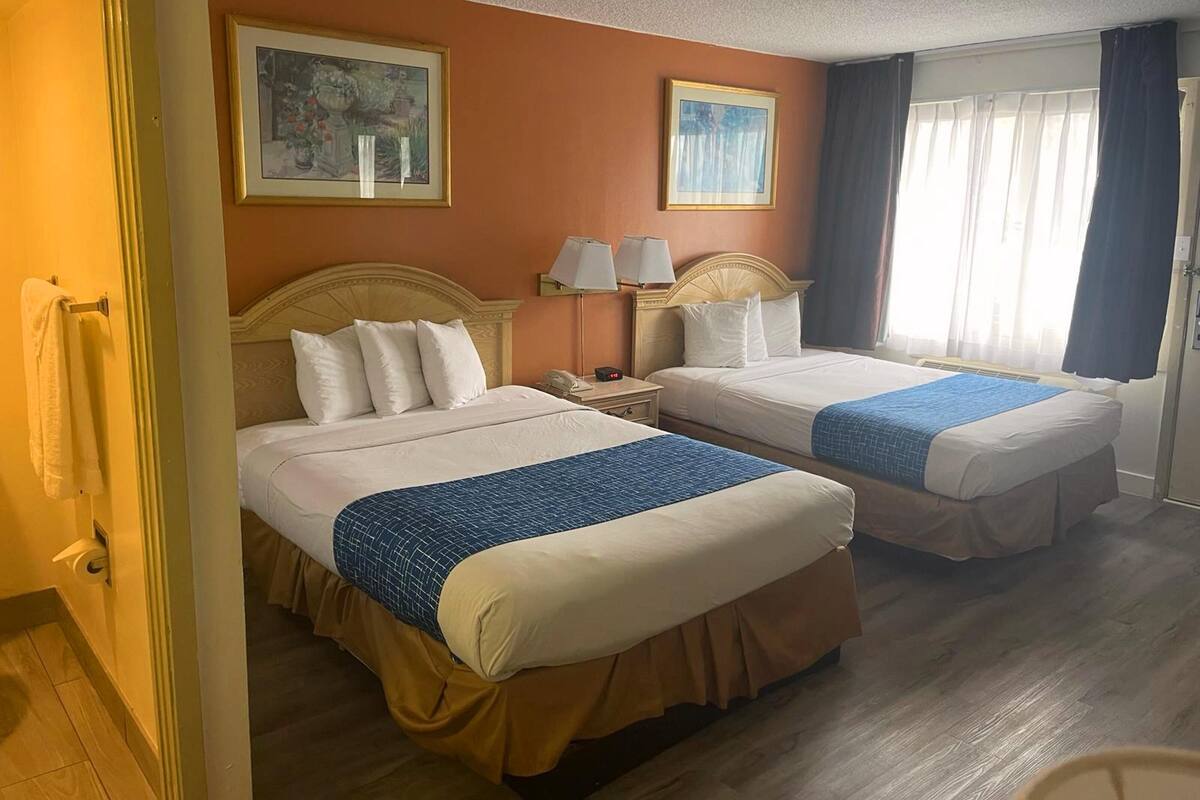 A hotel room with two beds, a wooden floor, a door with a towel rack, and two framed pictures on the wall.