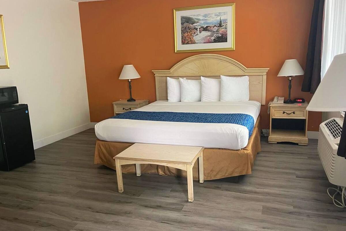 A hotel room with a large bed, two bedside tables, a small bedside table, a desk, a chair, a heater, and a picture on the wall.
