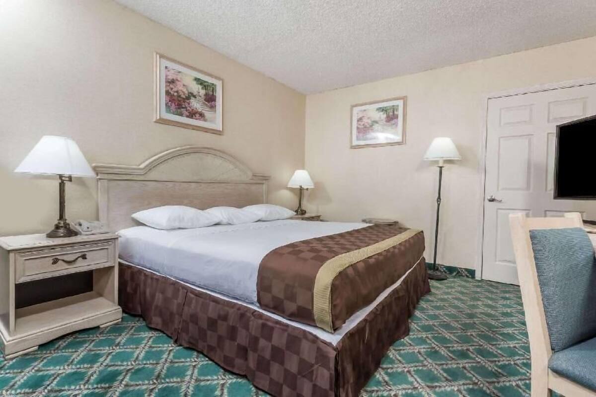 A hotel room with a bed, two bedside tables with lamps, a chair, a television, and two framed pictures on the walls.