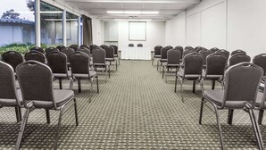 Un salón de conferencias con filas de sillas mirando una pantalla de proyección, suelo alfombrado y ventanas amplias.