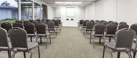 A conference room with rows of chairs facing a projection screen, carpeted floor, and large windows.