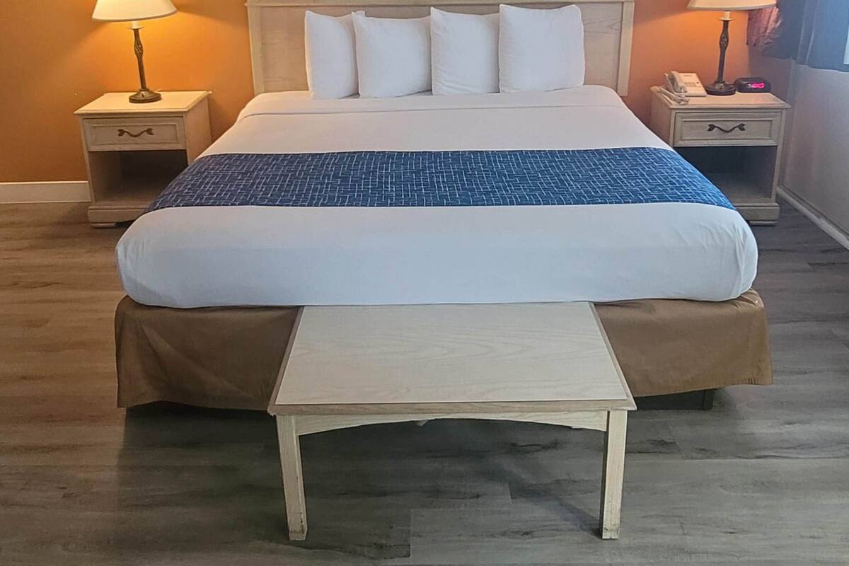 A neatly made bed with a blue and white bedspread, two bedside lamps, a framed picture on the wall, and a wooden bench in front of the bed.