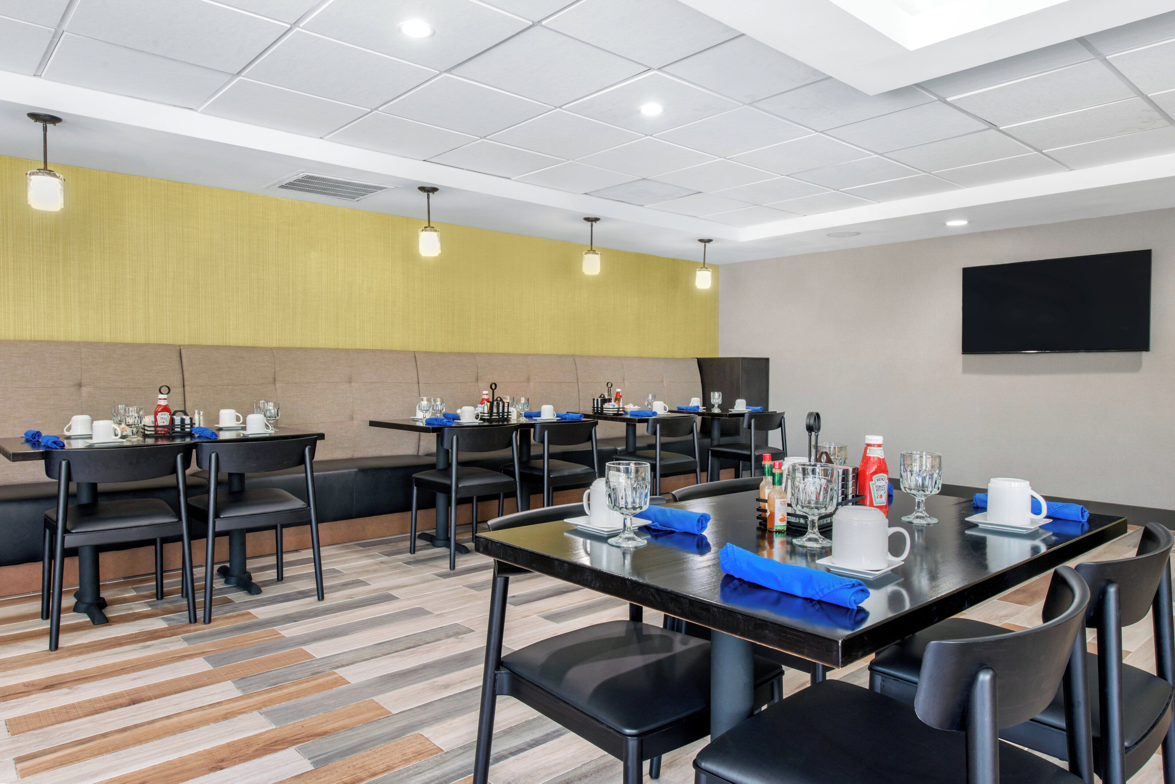 A hotel dining area with multiple tables, chairs, and a flat-screen TV mounted on the wall.