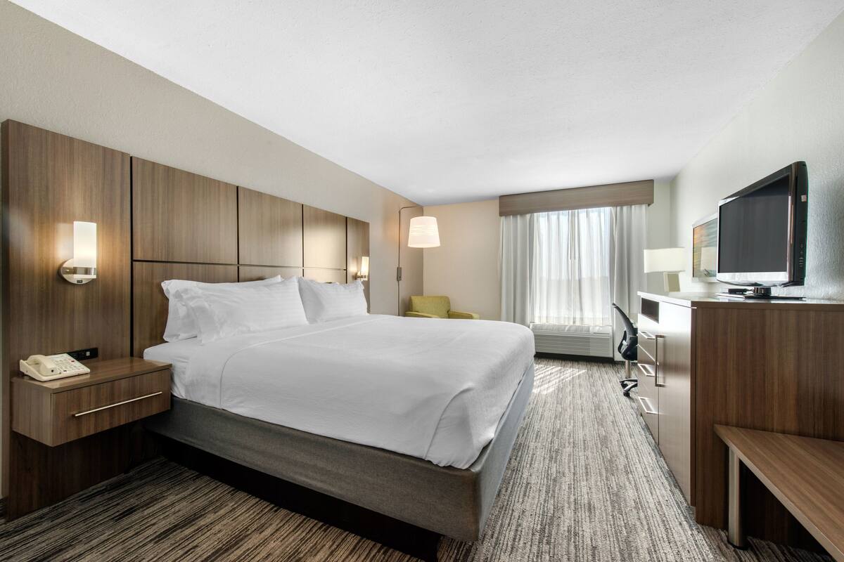 A hotel room with a large bed, bedside tables, a desk with a chair, a television, and a window with curtains.