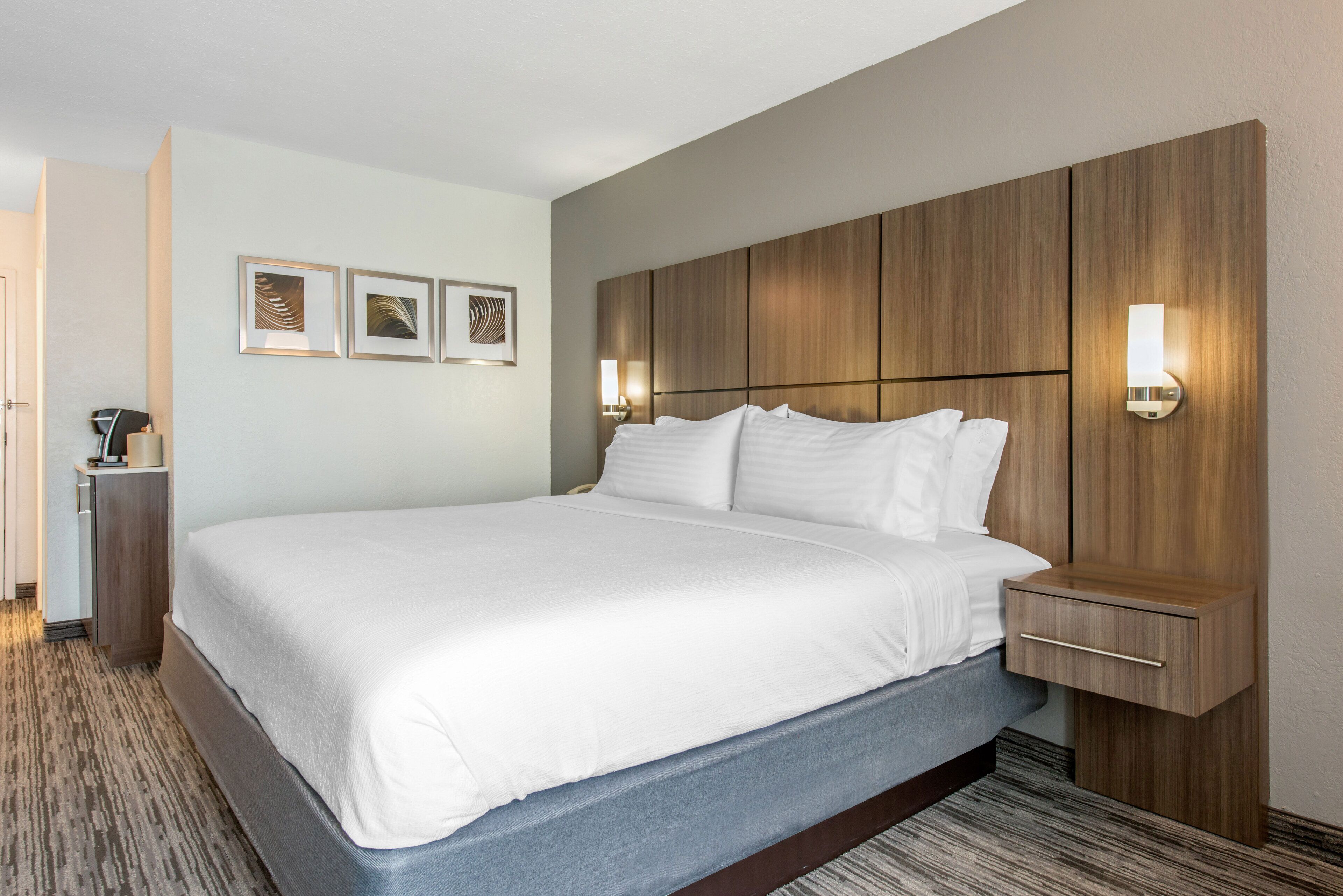 A hotel room with a large bed, wooden headboard, wall-mounted light fixtures, and framed artwork on the wall.