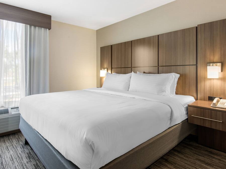 Une chambre d’hôtel avec un grand lit, une tête de lit en bois, des lampes de chevet, une table de chevet avec un téléphone et une fenêtre ornée de rideaux légers.