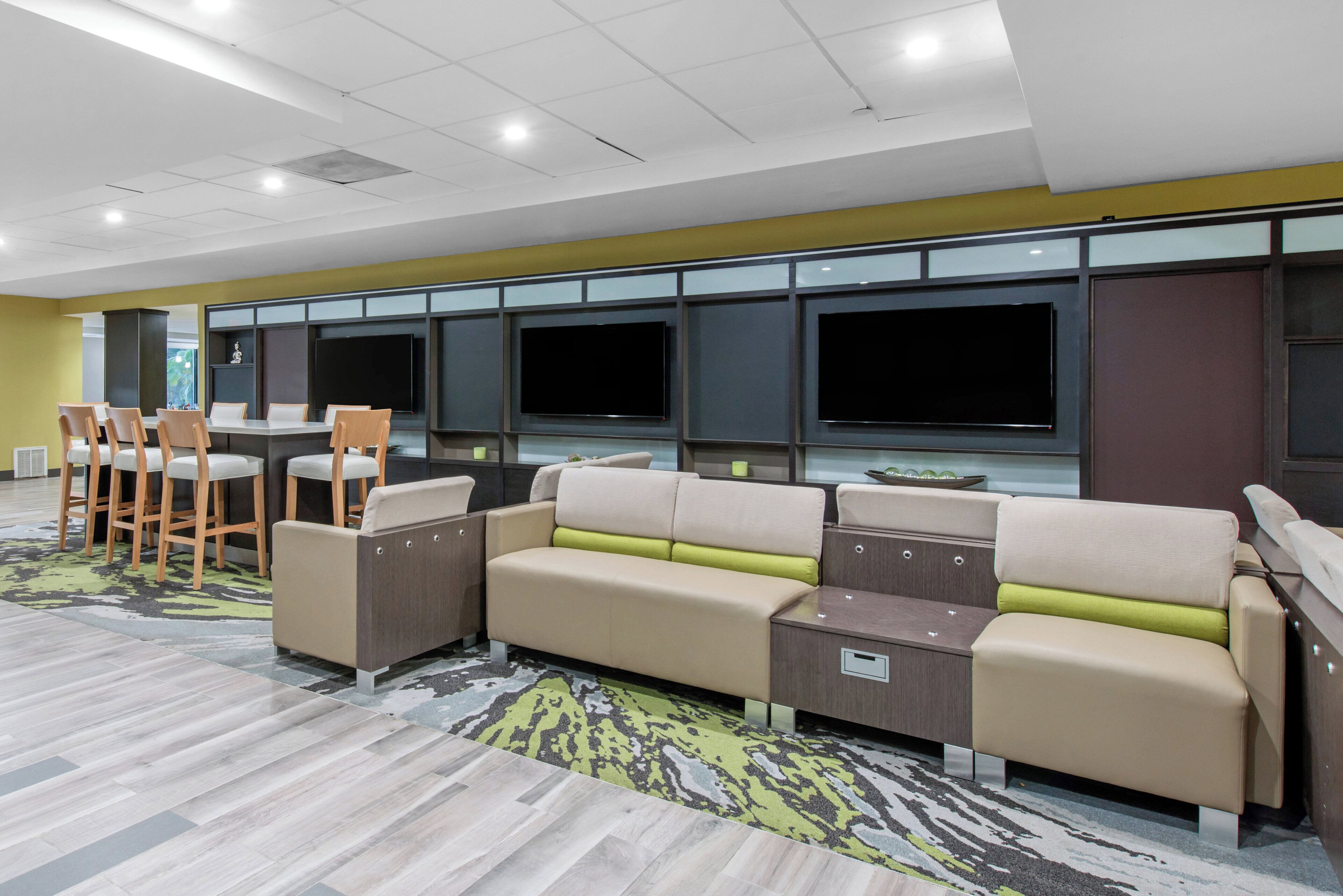 A hotel room with a sectional sofa, a small table, bar stools, and a flat-screen TV mounted on the wall.