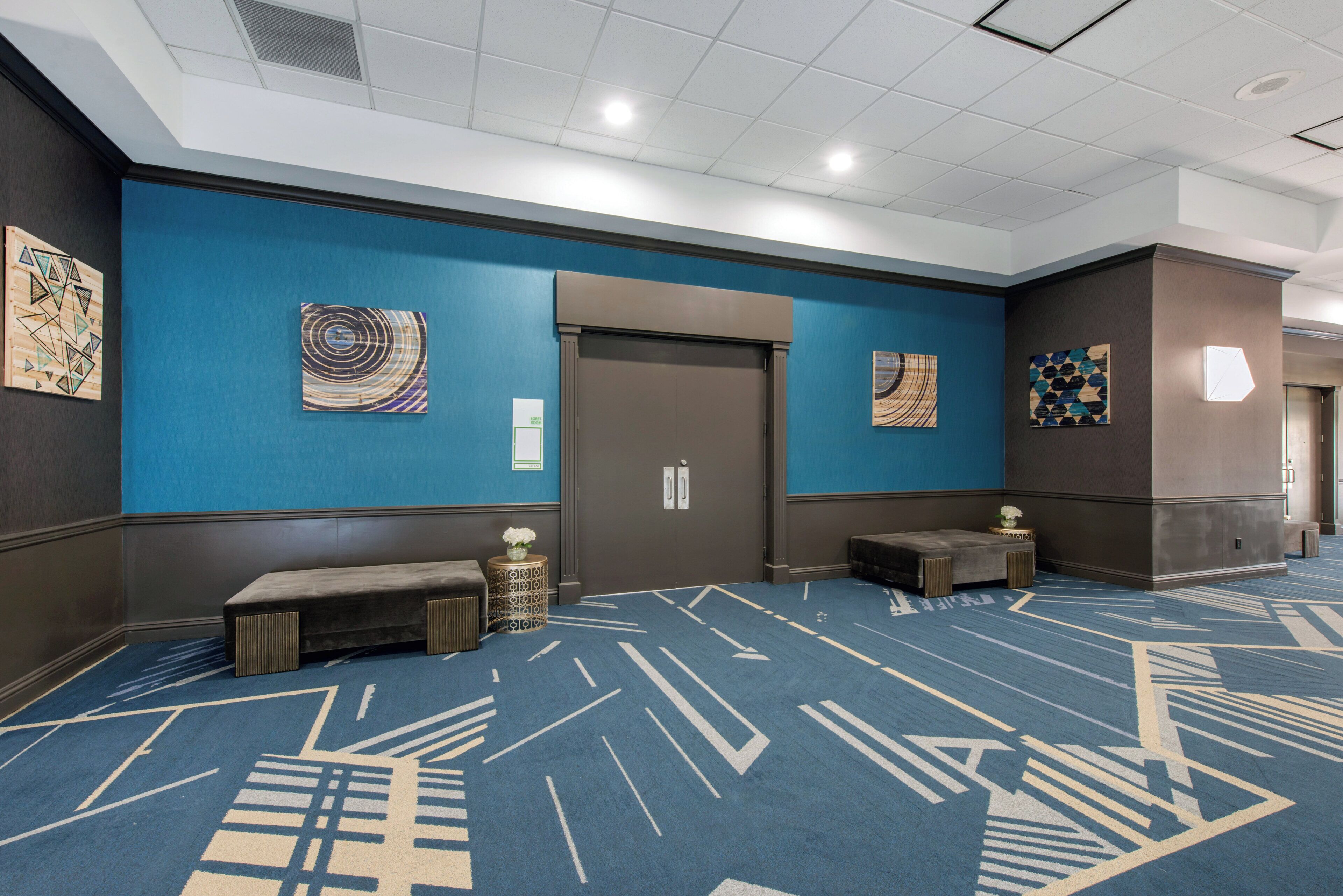 A hotel lobby with a blue carpet, artwork on the walls, a bench, and a door labeled 'EGRET ROOM'.