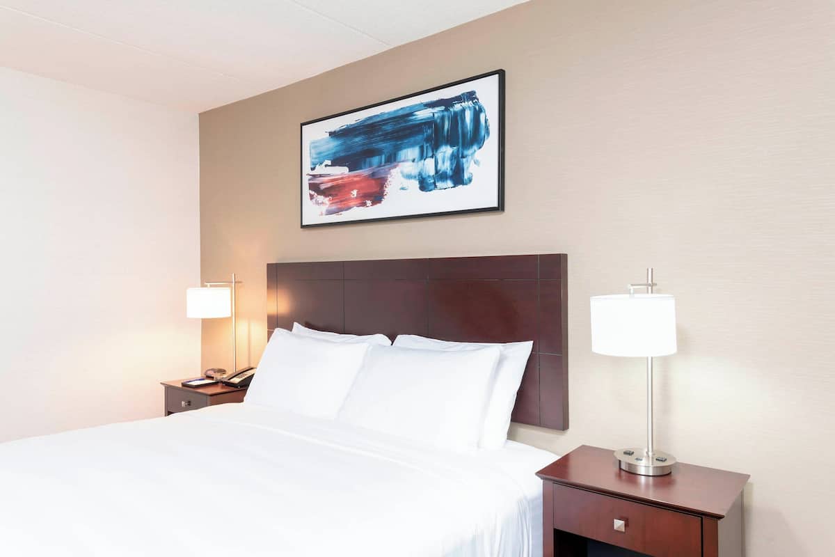 A hotel room with a large bed, a wooden headboard, a nightstand with a lamp, and a framed abstract painting on the wall.