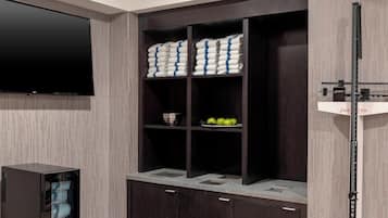 Una habitación moderna con un televisor de pantalla plana montado en la pared, un mueble de madera oscura con estantes que contienen toallas y un cuenco, y una balanza en el lado derecho.