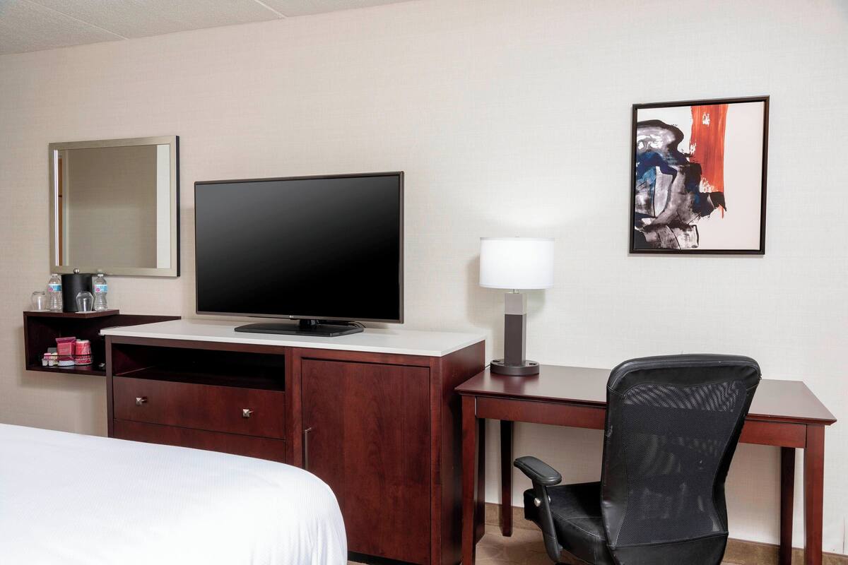 A hotel room with a bed, a TV, a desk with a chair, a lamp, and a framed picture on the wall.