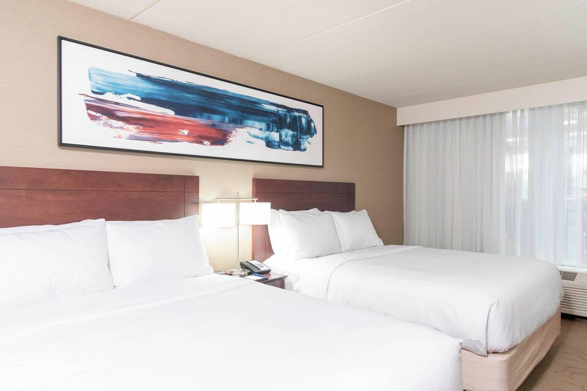 A hotel room with two beds, a large abstract painting on the wall, a bedside lamp, and a window with curtains.
