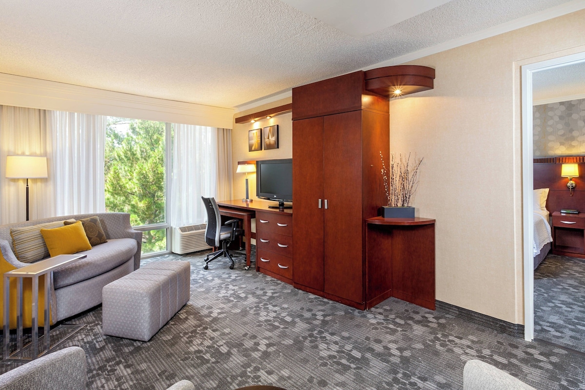 Une chambre d’hôtel avec un canapé gris, un bureau en bois, une télévision et une grande fenêtre avec des rideaux.