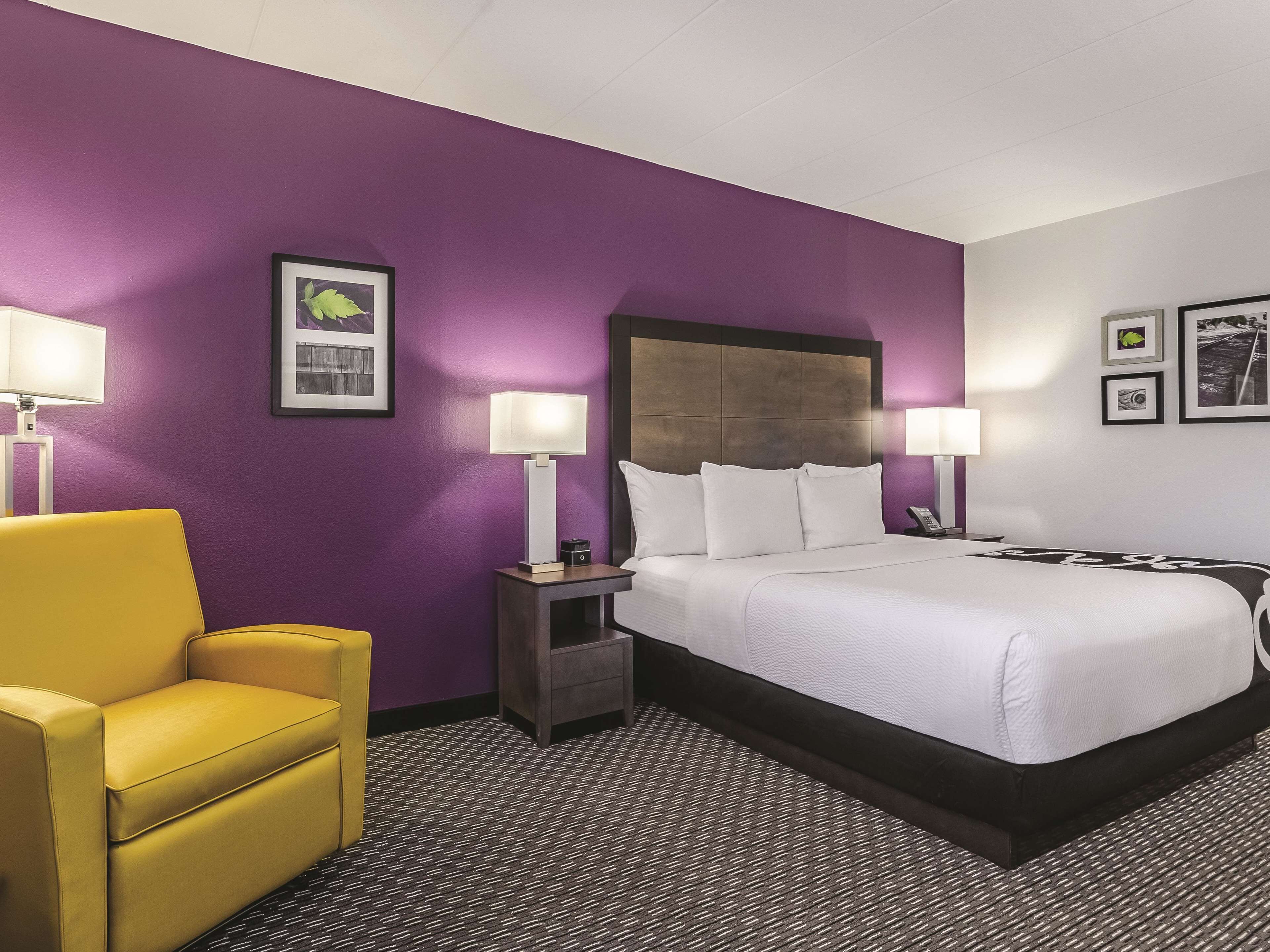 A hotel room with a large bed, two bedside tables with lamps, a yellow armchair, and a purple wall.