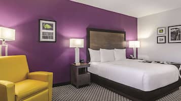 A hotel room with a large bed, two bedside tables with lamps, a yellow armchair, and a purple wall.