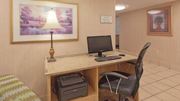 A hotel room with a desk, computer, printer, and a framed painting on the wall.