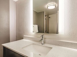 Un baño con encimera blanca con vetas, un lavamanos rectangular, un espejo y una ducha con barra de apoyo.