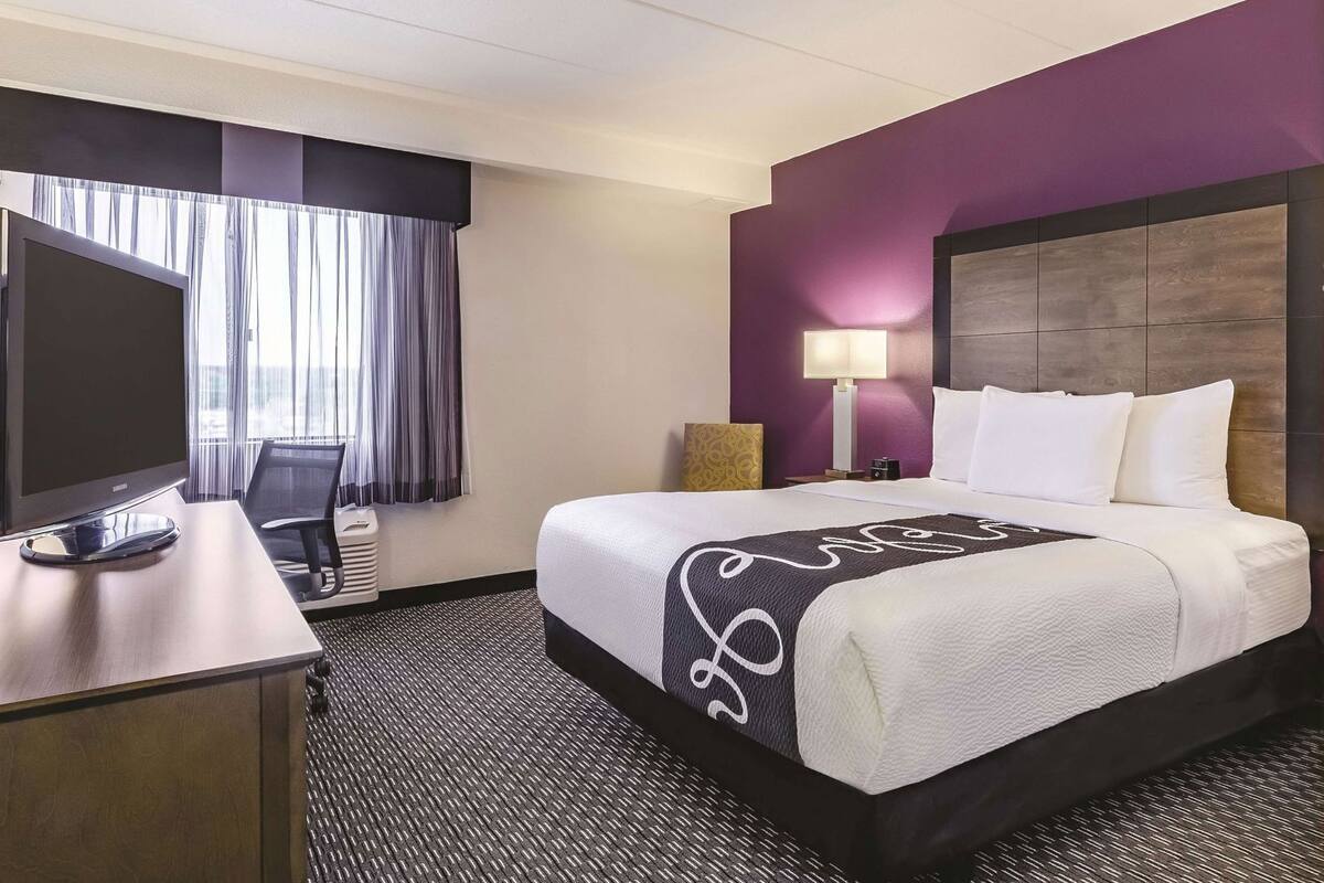 A hotel room with a bed, a desk with a TV, a lamp, and a window with curtains.