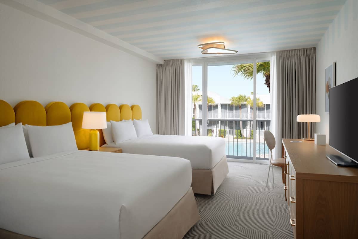 A hotel room with two beds, a desk, a TV, and a balcony with a view of palm trees.