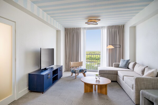 A modern hotel room with a flat-screen TV, a sofa, a chair, a coffee table, and a view of a green landscape through large windows.