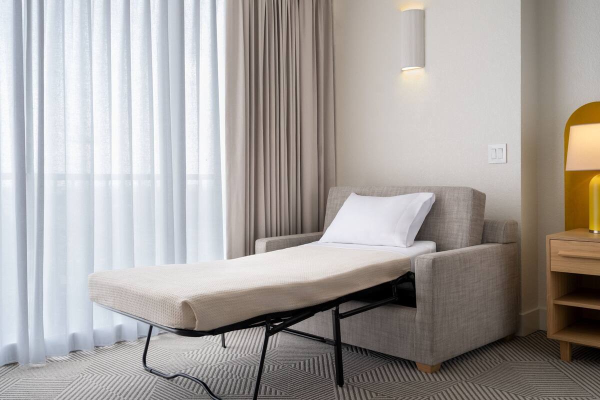A hotel room with a single bed, a bedside table, a lamp, and a window with curtains.