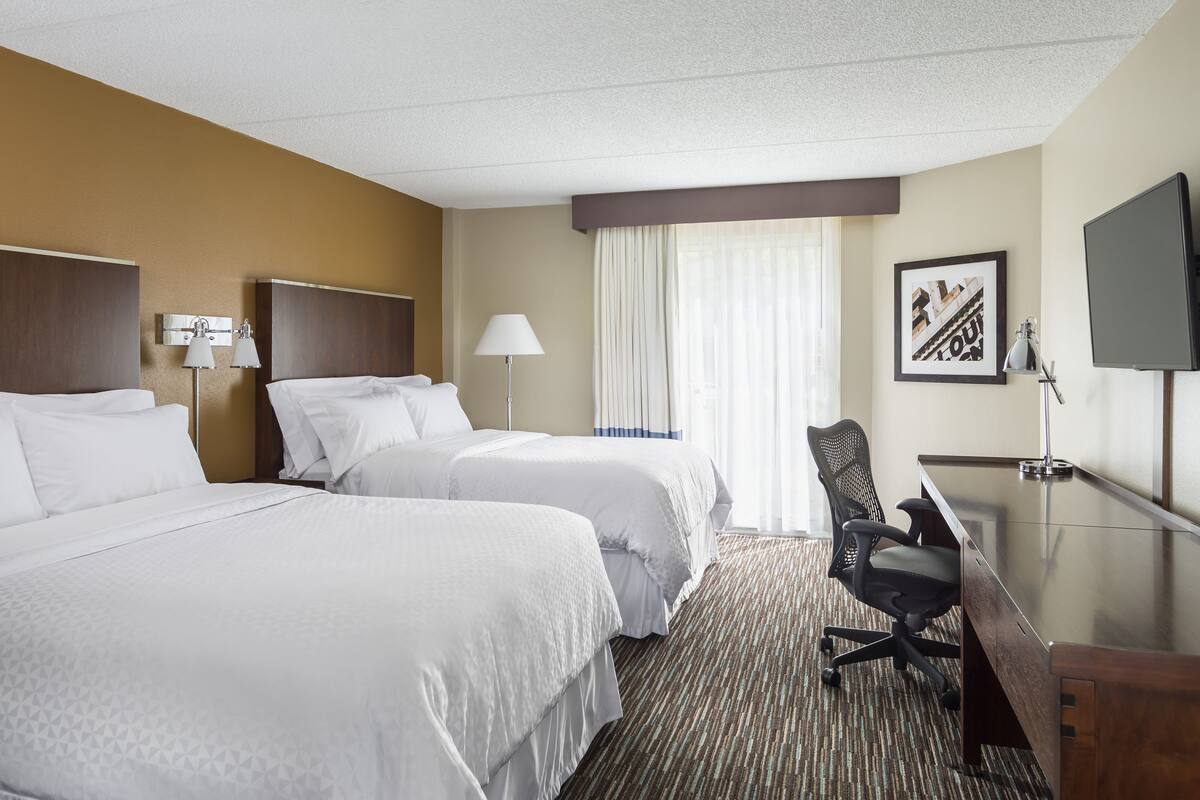 A hotel room with two beds, a desk, a chair, a TV, and a window with curtains.